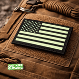 American flag morale patch with Velcro backing mounted on a backpack in daylight, showing black nylon fabric and green photoluminescent material.