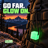 Green Mini Glow Cube hanging by a tied black paracord loop from a rugged hiking backpack overlooking a mountain lake at sunset.