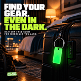 Bright green Forever Glow Tag with stainless steel cable loop attached to a black suitcase in a hotel room, making luggage easier to locate in darkness.