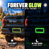 Green Forever Glow license plate frame on a black pickup truck in daylight and glowing green after dark.