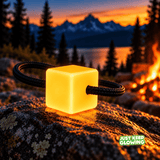 Single amber yellow Mini Glow Cube with black paracord threaded through its center, glowing on a rock at a mountain campsite.