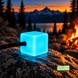 Single aqua blue Mini Glow Cube with a black paracord loop, glowing at a mountain campsite beside a campfire at sunset.