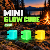 Three Mini Glow Cubes in aqua blue, green and amber yellow with black paracord, glowing on a rock beside a backpack and campfire at a mountain campsite.