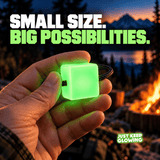 Hand holding a small green glow in the dark cube with black paracord at a campsite, demonstrating its compact 1.25 inch size.
