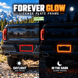 Red license plate frame on a black pickup truck during daylight and glowing orange in darkness.