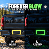 Yellow license plate frame installed on a black pickup truck in daylight and glowing green at night.